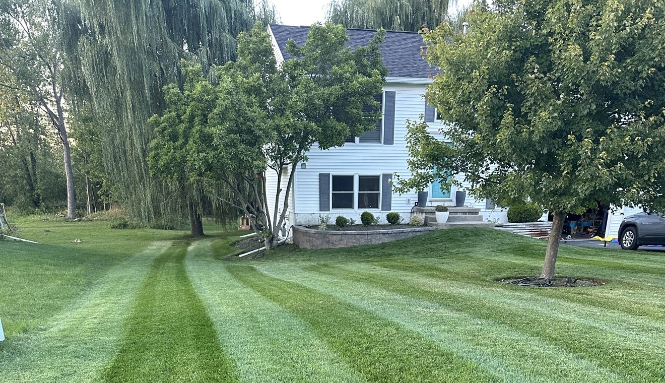 Freshly striped green lawn, the mowing stripes converging toward a white house — a Bordoy Outdoor Services mowing job